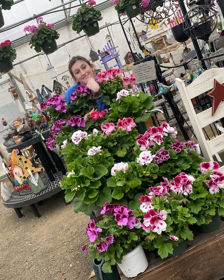 Woman smiling among vibrant geranium flowers in a greenhouse, surrounded by decorative garden items and hanging plants, showcasing a colorful floral display.