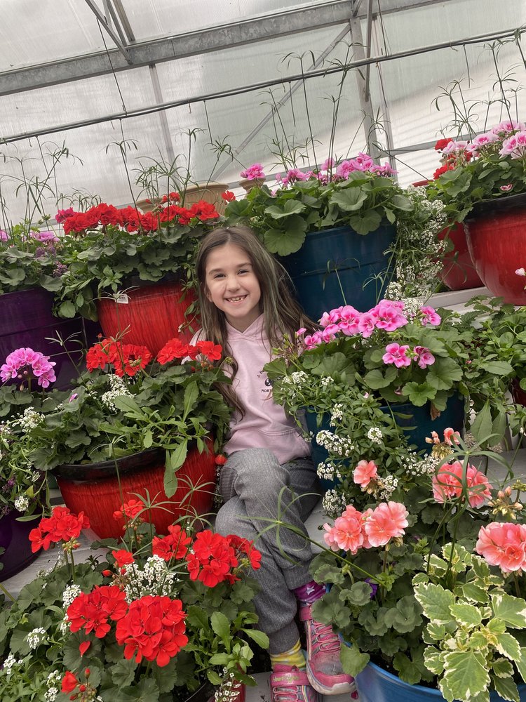A young girl smiles among vibrant flower pots in a greenhouse, surrounded by colorful geraniums and other blooming plants, showcasing the beauty of gardening and nature.