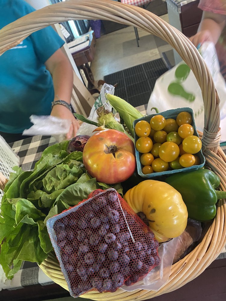 A wicker basket filled with fresh produce including lettuce, apples, yellow and green tomatoes, a yellow squash, green bell pepper, and a bag of blueberries, with a blurred background of a market setting.