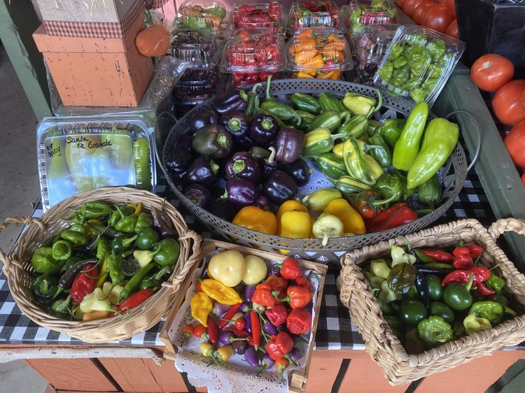 Colorful assortment of fresh vegetables displayed at a market, featuring various types of peppers including green, red, yellow, and purple. Baskets and trays showcase the vibrant produce, highlighting organic farming and seasonal offerings.