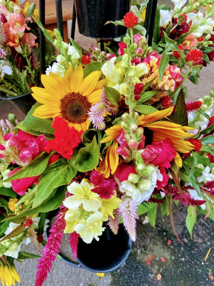 Vibrant bouquet featuring sunflowers, snapdragons, and various colorful flowers arranged in a black pot, perfect for enhancing outdoor decor or special occasions.