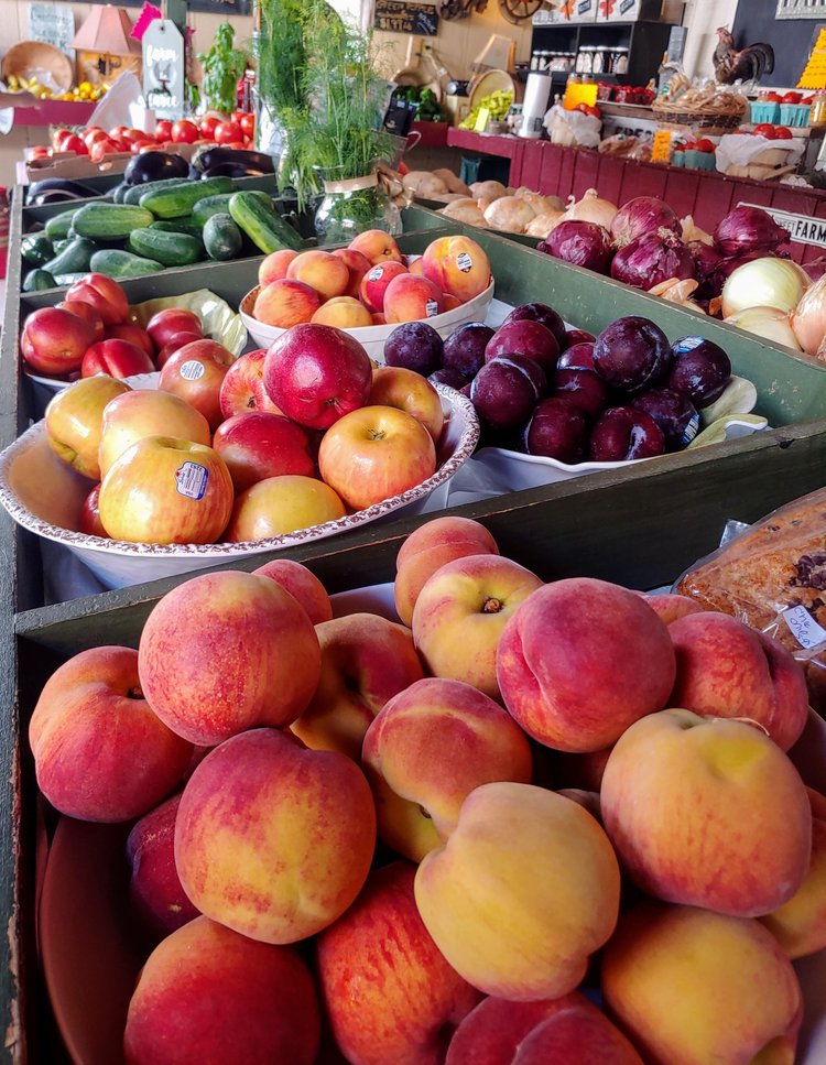 Fresh fruits and vegetables displayed at a local farmers market, featuring vibrant peaches, apples, plums, cucumbers, and various baked goods.